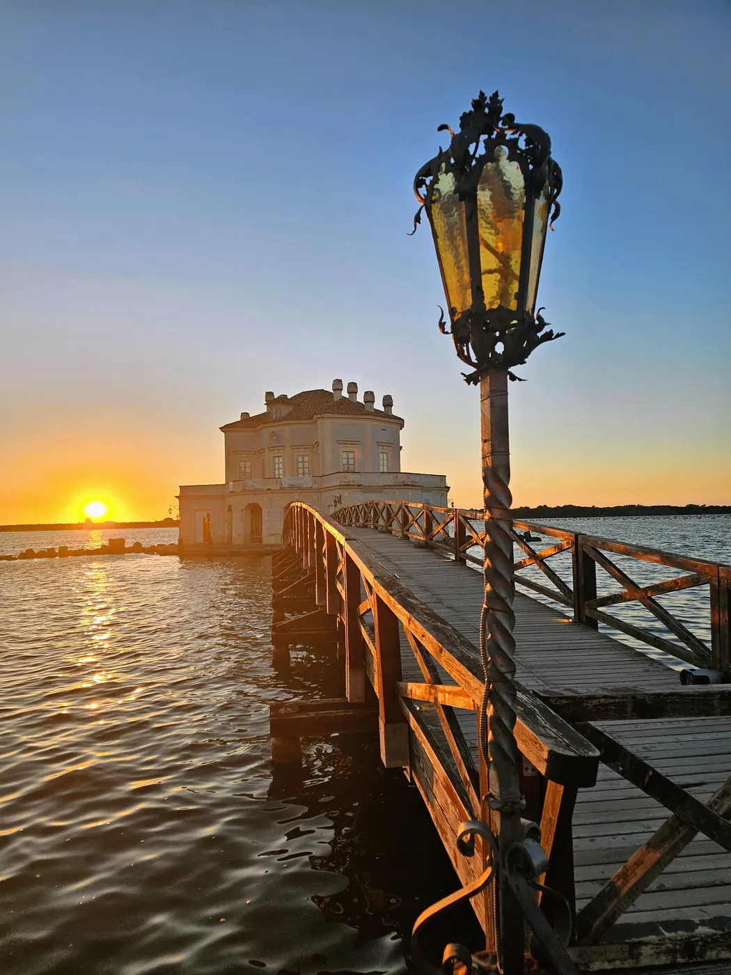 La Casina Vanvitelliana sul lago Fusaro al tramonto, con il pontile di legno e un lampione in primo piano