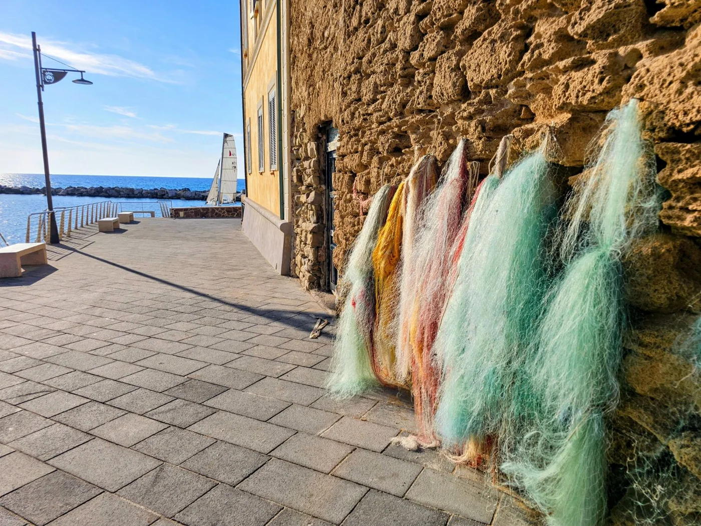 Reti da pesca verdi, gialle e rosse appese a un muro di pietra sul lungomare di Santa Maria di Castellabate, con il mare e una vela sullo sfondo.