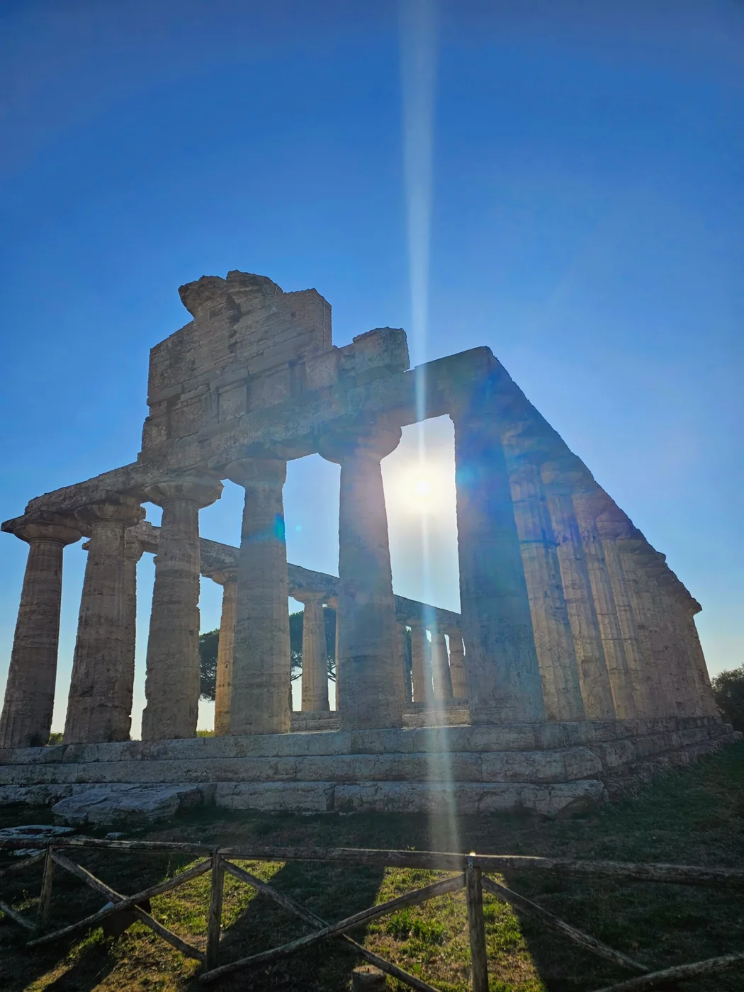 Il tempio di Atena a Paestum visto in controluce, con il sole che filtra fra le colonne doriche contro un cielo blu intenso.