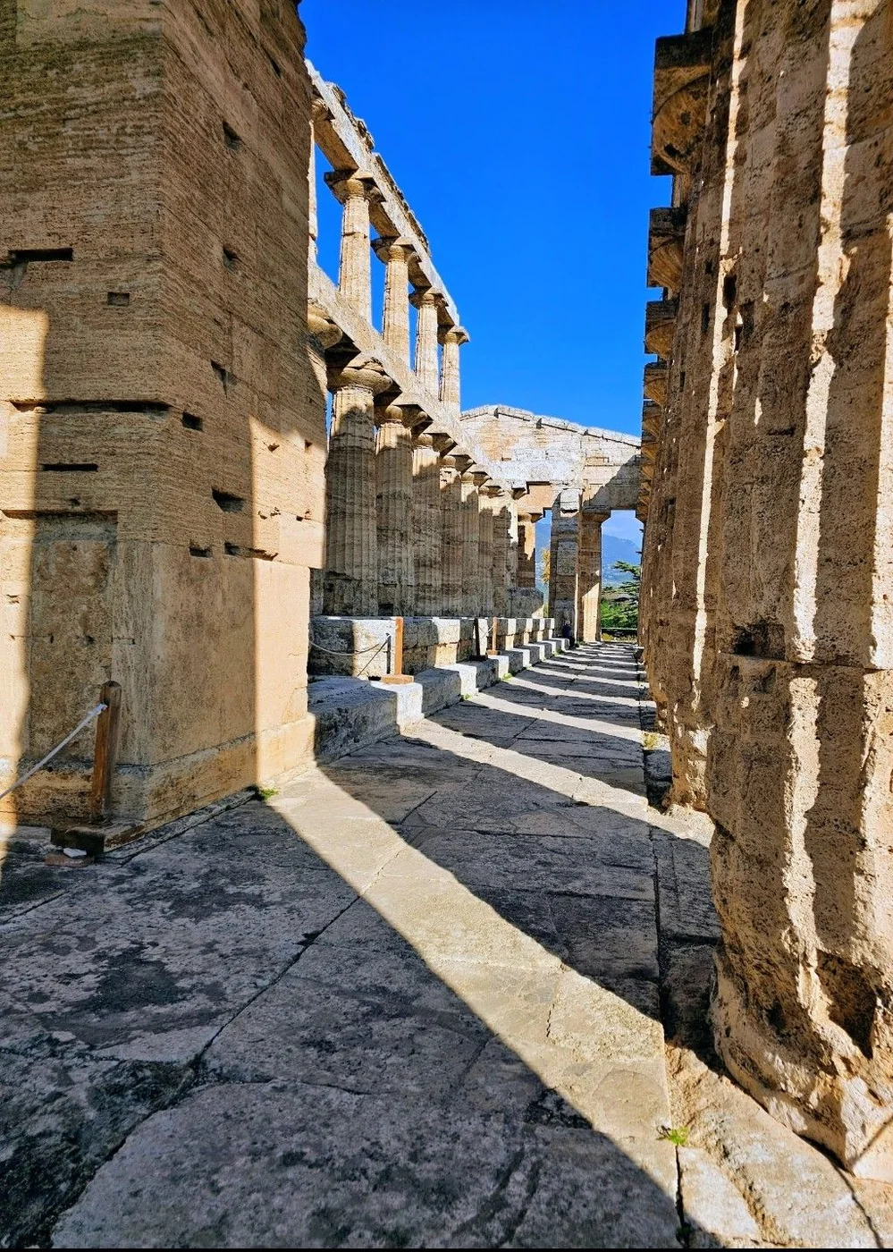 Un passaggio fra i massicci blocchi del tempio di Nettuno a Paestum, con le ombre delle colonne sul pavimento e il cielo blu in fondo.