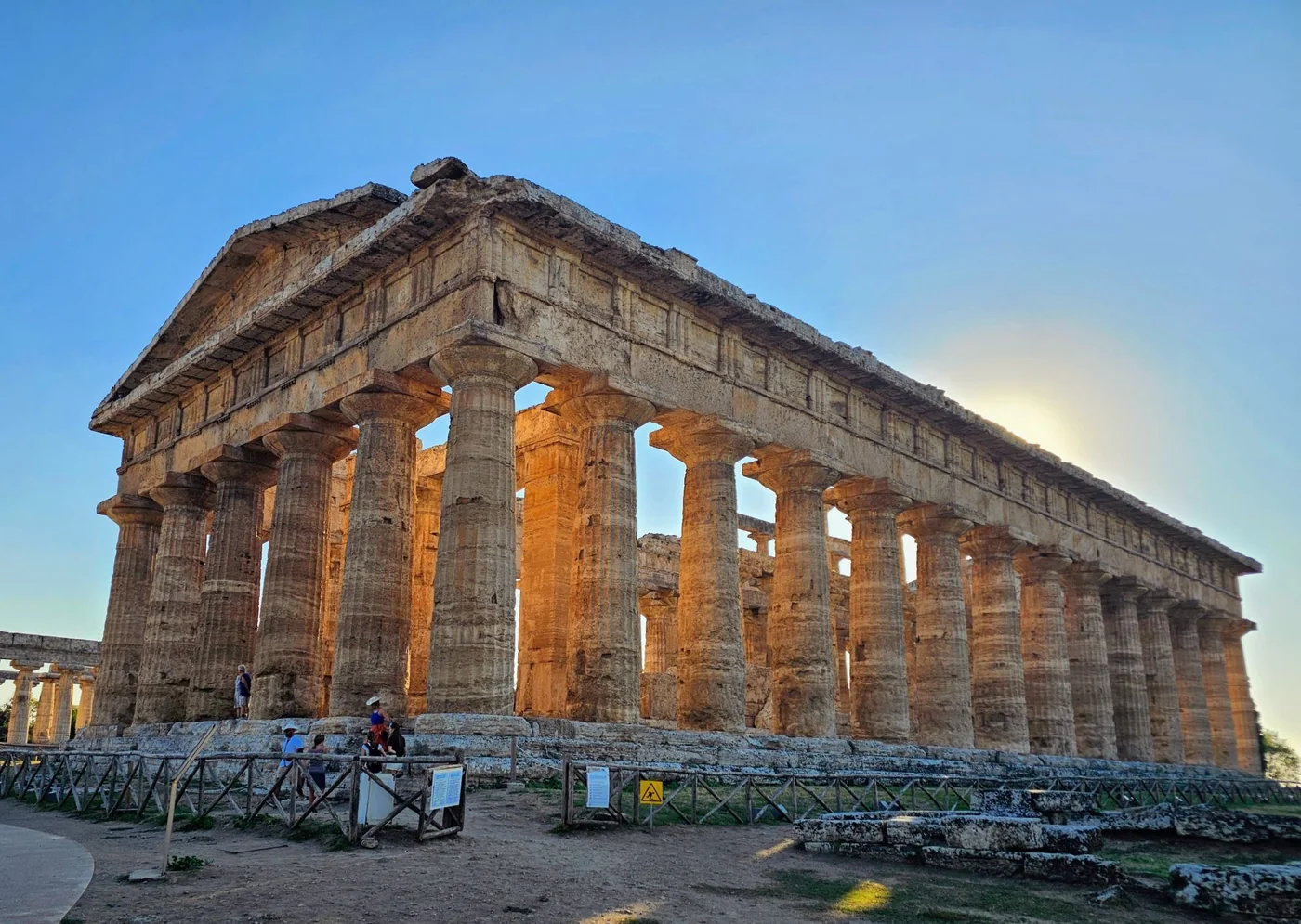 Il tempio dorico di Nettuno a Paestum nella luce arancione del tardo pomeriggio, con le colonne di travertino e i visitatori ai piedi del basamento.