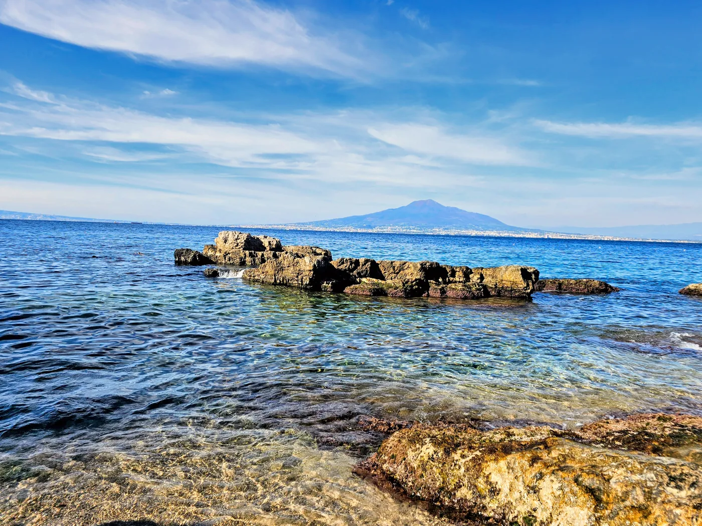 Scogli nell'acqua limpida e, all'orizzonte, il Vesuvio oltre il golfo di Napoli
