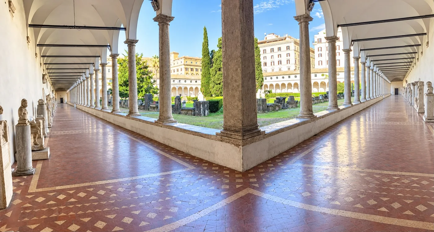 Il chiostro di Michelangelo alle Terme di Diocleziano a Roma, con i portici ad arcate, le colonne di travertino, i cipressi nel giardino e le statue lungo il pavimento di cotto.