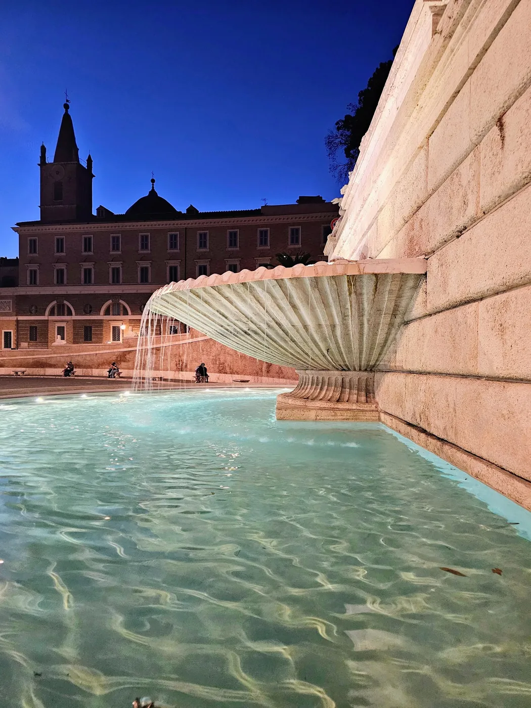 L'acqua scende da una grande vasca a conchiglia in una fontana addossata al muro di piazza del Popolo, all'imbrunire, con una chiesa e i palazzi della piazza sullo sfondo