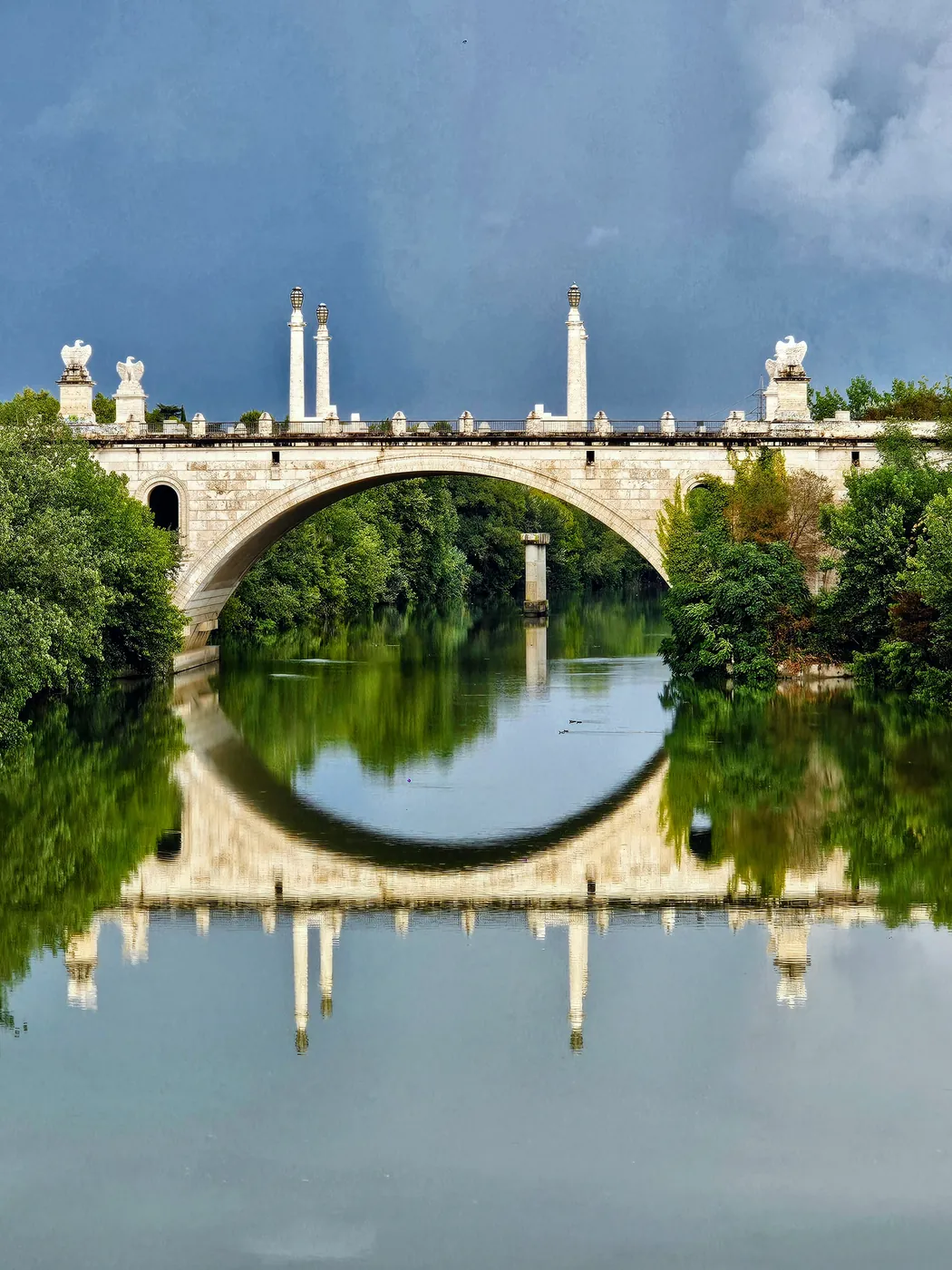 L'arcata di Ponte Milvio con le statue in cima, riflessa nell'acqua ferma del Tevere tra gli alberi