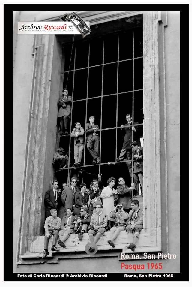 Fotografi e curiosi affacciati e seduti su una grande finestra con inferriata che dà su piazza San Pietro