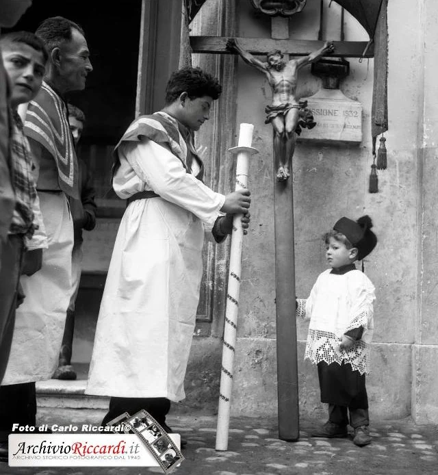 Un chierichetto regge un grosso cero durante una processione; accanto al crocifisso un bambino in cotta e cappello lo guarda